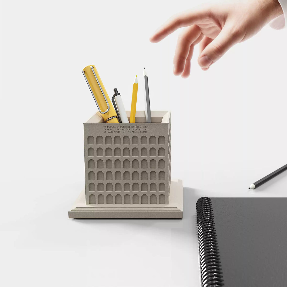 A hand reaching toward stationery in the Square Colosseum pen holder on a desk.